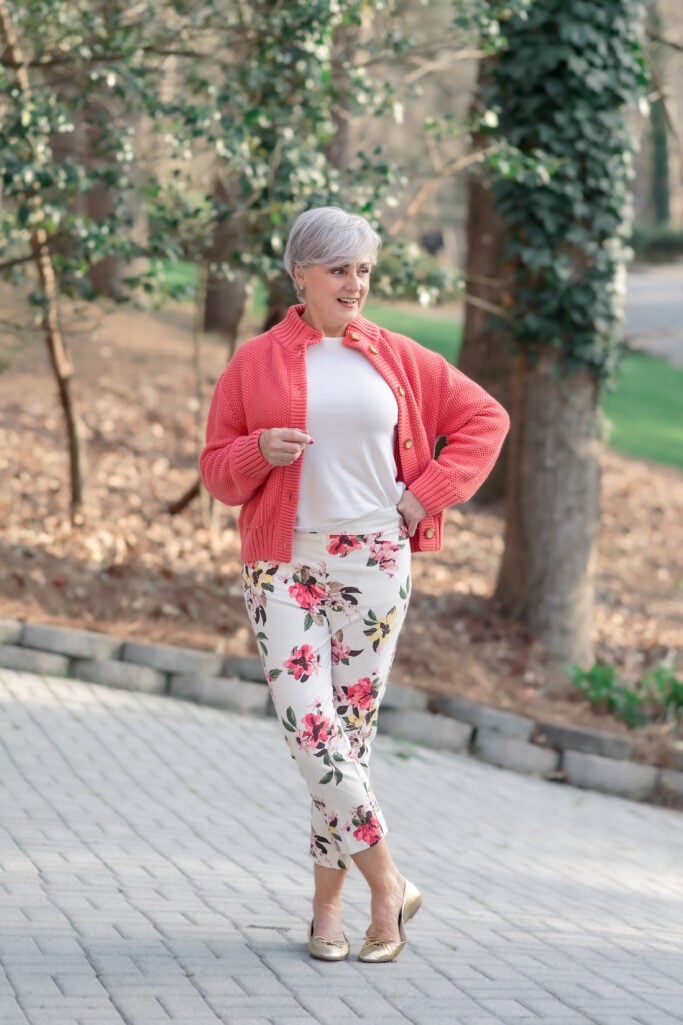 Woman over 50 with silver hair wearing a coral chunky-knit button-front sweater jacket over a white fitted cotton tee, paired with cropped floral slim pants in cream with pink and green blooms, and gold metallic ballet flats — a warm, spring-ready coral look for women over 50 that coordinates color through print.