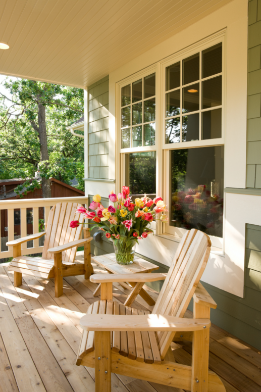 Cozy front porch with wooden Adirondack chairs and a small table holding a vibrant bouquet of tulips, bathed in warm natural light with lush greenery in the background.