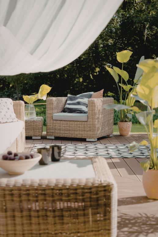 Outdoor patio lounge with woven wicker chairs, neutral cushions, and a patterned rug, styled with potted plants and soft white drapery for a relaxed, sunlit backyard setting.