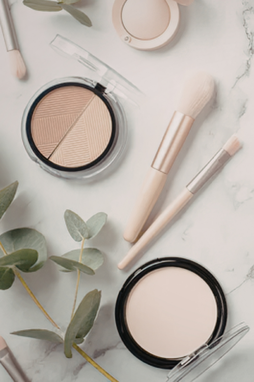 Flat lay of neutral-toned makeup including pressed powders and soft-bristle brushes, styled with greenery on a light marble surface for a clean, natural beauty aesthetic.