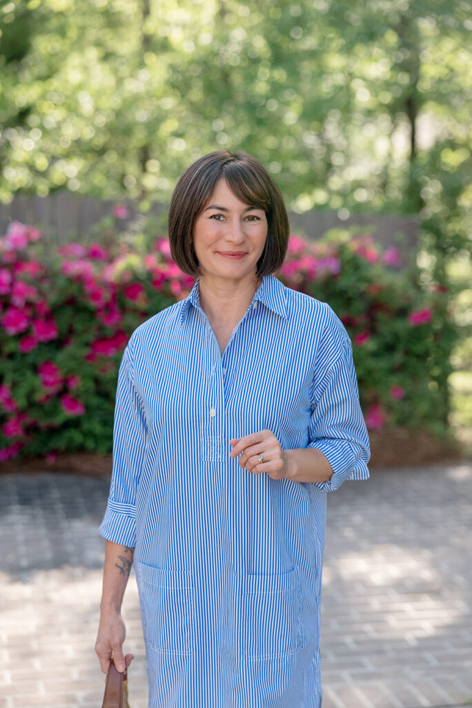 Kelly smiling in blue and white striped broadcloth shirtdress with rolled sleeves, holding straw hobo bag, pink azaleas in background — petite shirtdress spring outfit