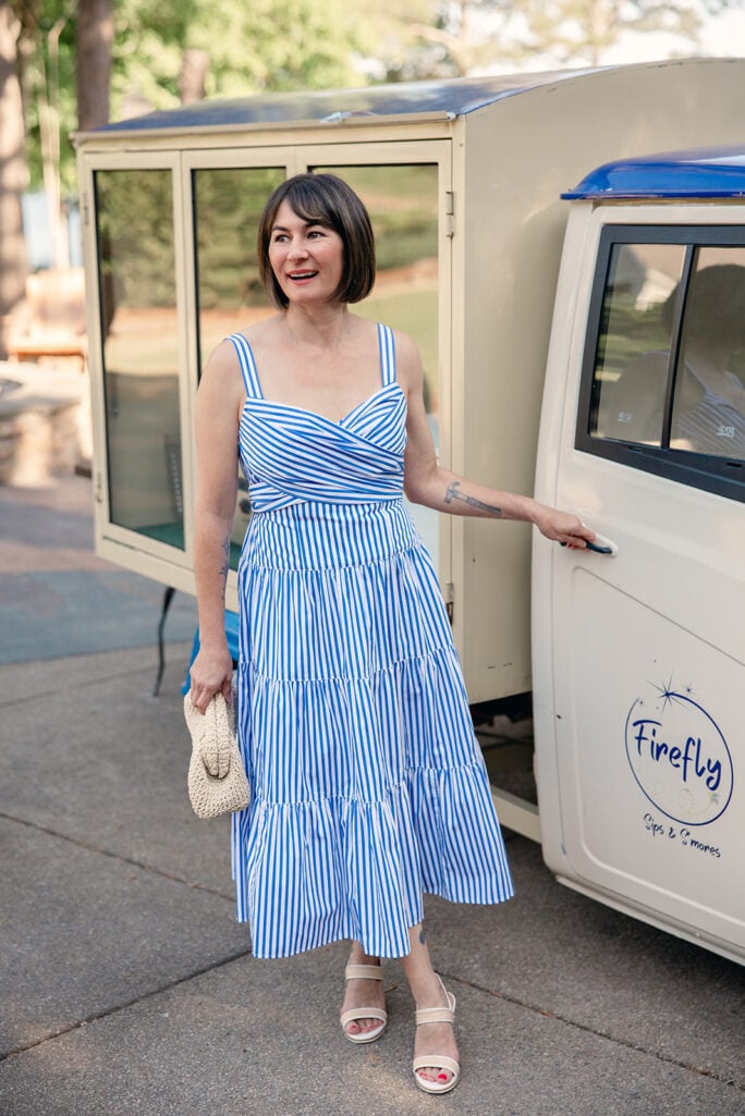 Kelly smiling in blue and white striped cotton tiered dress with crossover bodice and back bow, holding straw clutch with nude wedge sandals — tiered dress for petites