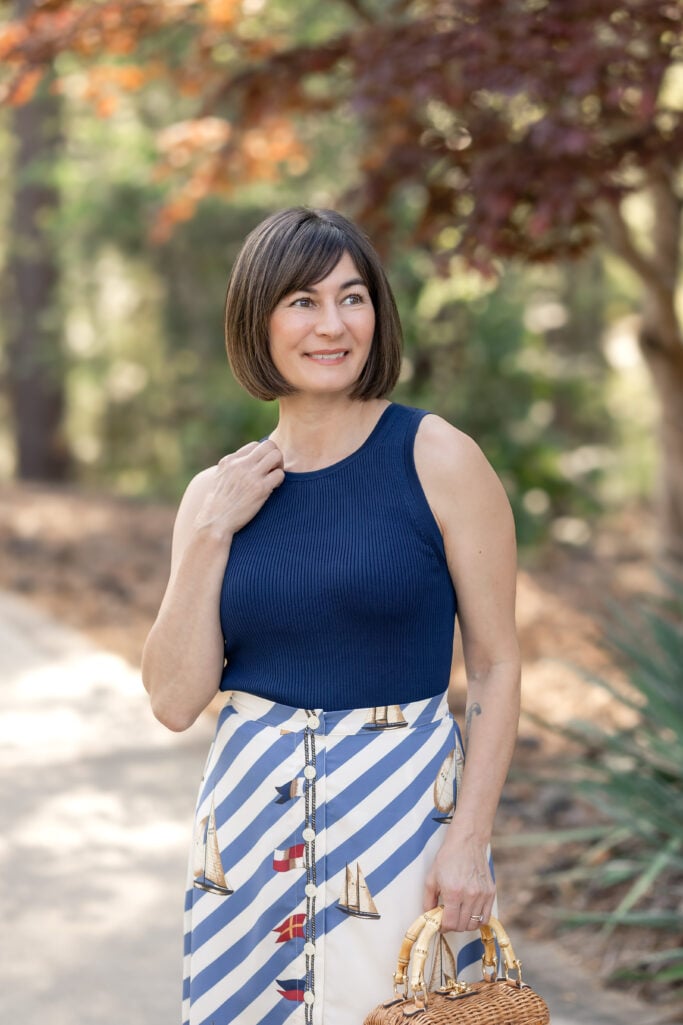 Three-quarter shot of Kelly in navy ribbed knit top tucked into Ralph Lauren sailboat stripe midi skirt with wicker bamboo bag — nautical petite outfit spring