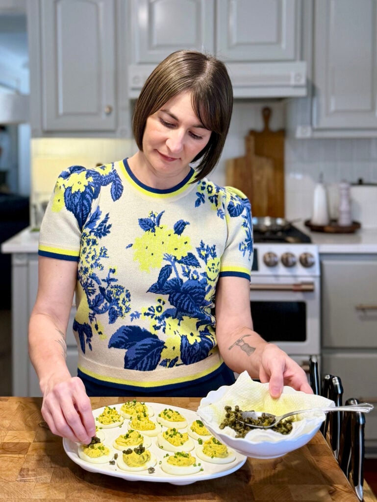 Kelly stands at a butcher block island in the kitchen, carefully topping a platter of deviled eggs with crispy capers from a small bowl, wearing her blue and yellow floral sweater tee.