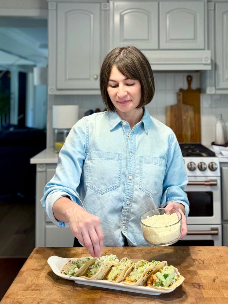 Kelly stands at the butcher block island in the kitchen, sprinkling crumbled cotija cheese from a glass bowl over a white rectangular platter of assembled grilled shrimp tacos topped with avocado crema and slaw, wearing her light chambray cropped top.