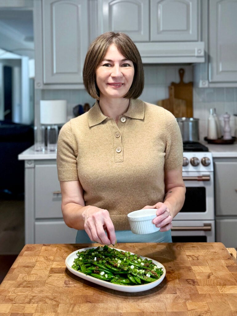 Kelly stands at the butcher block island in the kitchen, finishing a platter of sugar snap peas with sesame ginger glaze, sprinkling green onions from a small white ramekin, smiling at the camera in her camel polo sweater.