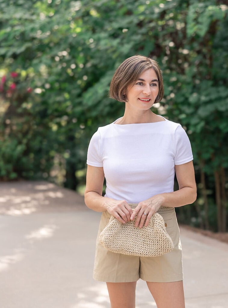 Woman with short brown bob hair wearing a white boat-neck short-sleeve tee with khaki tailored shorts, holding a natural crocheted raffia dumpling-style clutch bag with a pleated top and soft rounded silhouette, walking on a paved path with green foliage behind her — a minimalist raffia clutch styled casually for spring.
