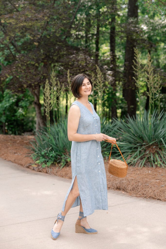 Woman with dark bob hair walking on a sidewalk wearing a chambray blue sleeveless linen button-front midi dress with side slits, carrying a round natural rattan basket bag with tan leather top handles, and wearing chambray blue ankle-tie espadrille wedges with jute-wrapped heels — a classic spring raffia wedge and basket bag pairing over 50.