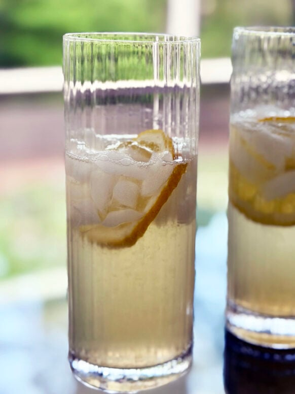 Two Elderflower Collins cocktails in tall ribbed highball glasses sit on a glass table, each garnished with a lemon twist and ice, glowing pale golden in the spring light with soft green bokeh in the background.