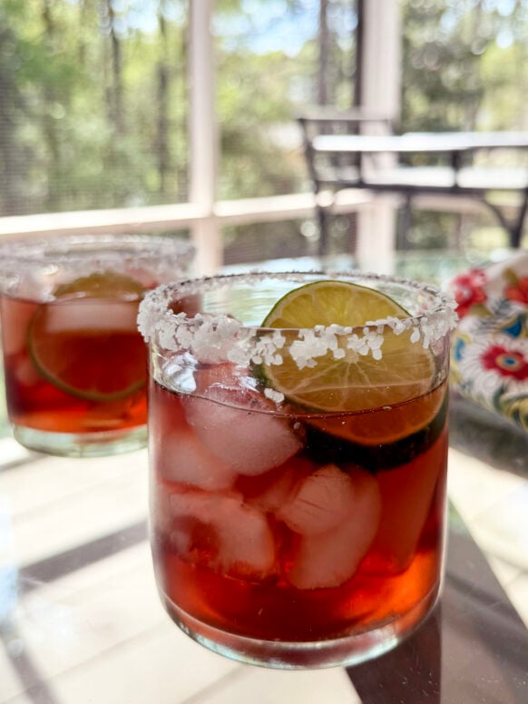 Two Hibiscus Margaritas in rocks glasses with salted rims and lime wheel garnishes sit on a glass table on the screened porch, glowing deep ruby red in the spring light, a floral linen visible in the background.
