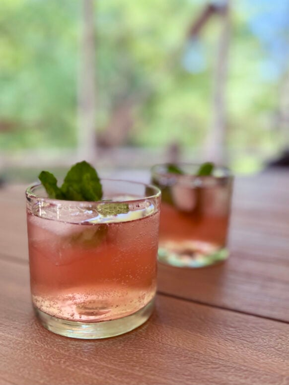 Two rocks glasses filled with sparkling rosy pink berry sangria, each garnished with a fresh mint sprig, sitting on a weathered wooden outdoor table with soft green bokeh background.