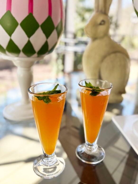 Two Peach Bellinis in classic champagne flutes sit on a glass table garnished with fresh mint sprigs, a ceramic Easter bunny and pink and green decorative eggs visible in the soft background.