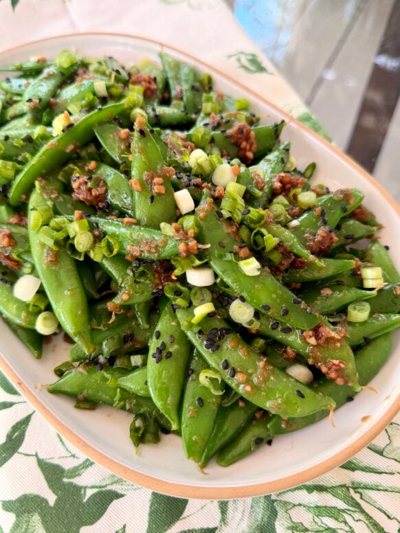 A generous oval platter of bright green sugar snap peas tossed in a sesame ginger glaze, topped with sliced green onions and black sesame seeds, set on a green and white botanical linen.