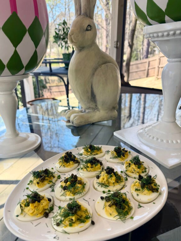 A white oval platter of deviled eggs topped with fresh tarragon, chives, and crispy capers sits on a glass table, with a ceramic Easter bunny and decorative pink and green Easter eggs in the background.