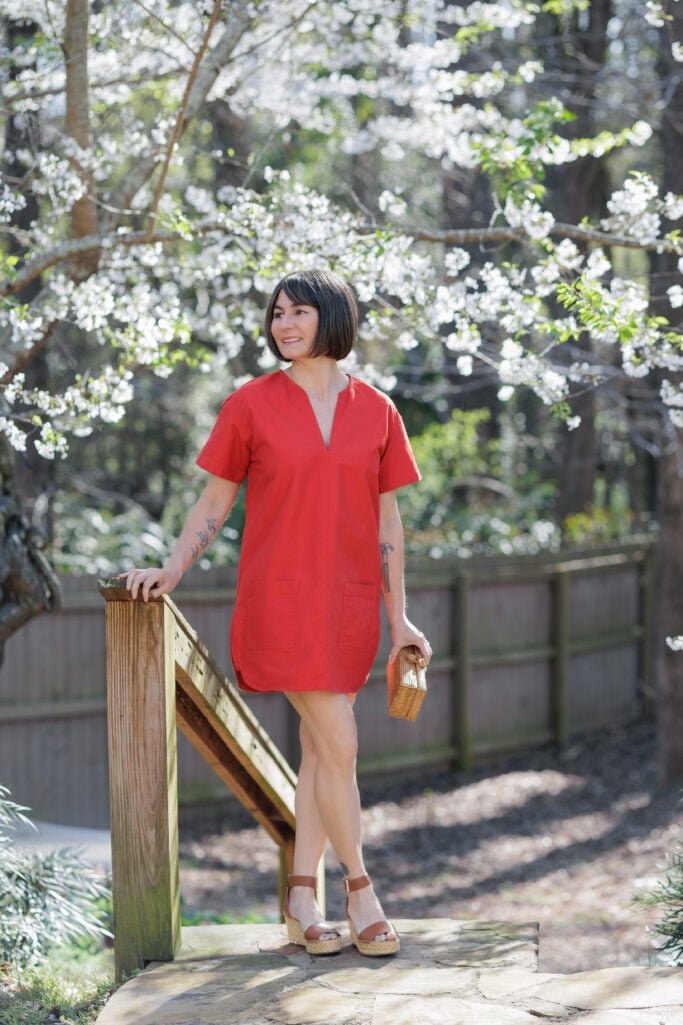 Kelly in red shift dress with Franco Sarto raffia wedge sandals and straw clutch standing on stone garden steps, cherry blossoms — full length petite spring outfit