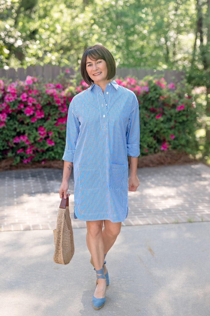 Kelly smiling in blue striped broadcloth shirtdress with Castañer espadrille wedges and straw hobo bag, pink flowers in background — petite shirtdress summer look
