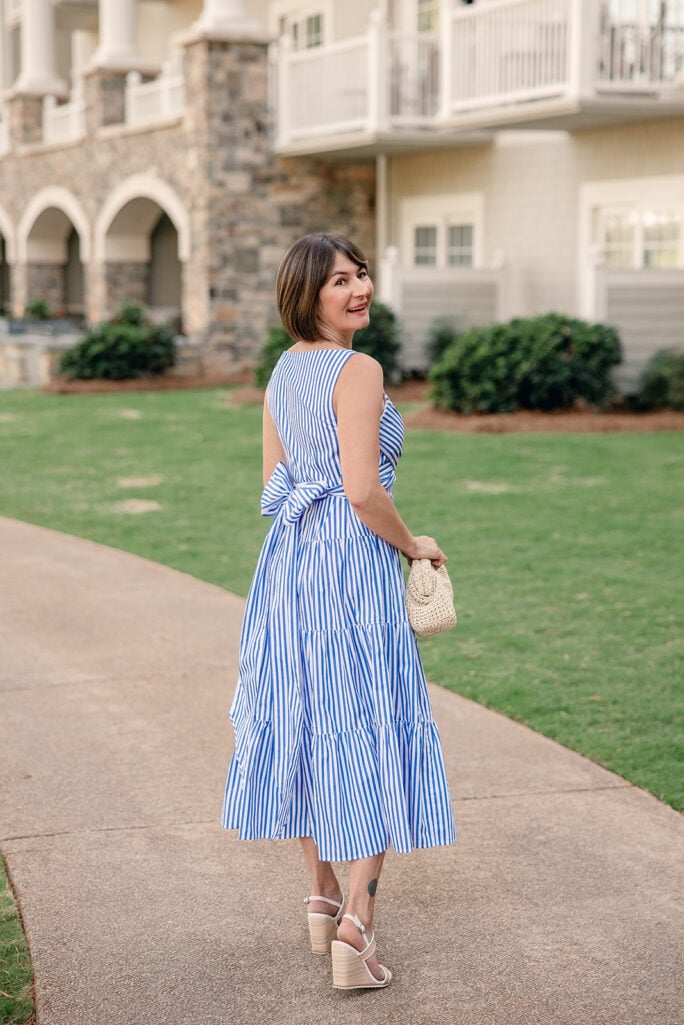 Full-length back view of Kelly in striped tiered cotton dress showing bow waist detail, tiered skirt volume and nude wedge sandals — tiered dress petite silhouette