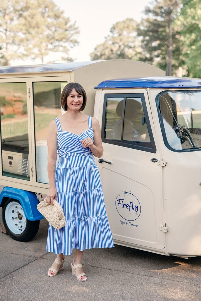 Kelly smiling in striped tiered midi dress with crossover bodice beside vintage truck, straw clutch in hand — petite occasion dress summer outfit