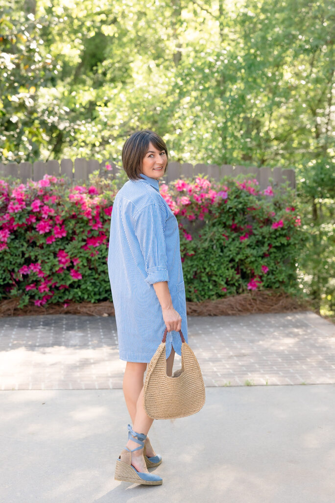 Back view of Kelly in blue and white striped shirtdress with straw hobo bag and blue lace-up espadrille wedges, pink azalea backdrop — petite shirtdress back silhouette