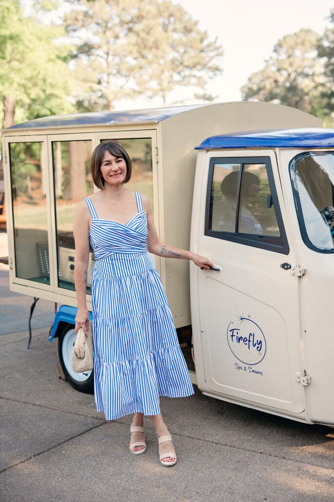 Kelly smiling in striped tiered midi dress with crossover bodice beside vintage truck, straw clutch in hand — petite occasion dress summer outfit