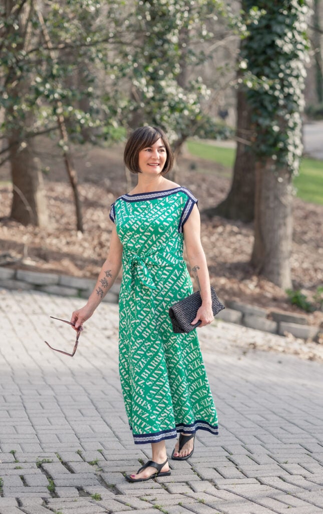 Woman wearing a bold kelly green and ivory typography print midi dress with navy trim at the neckline and hem, a self-tie waist detail, black thong sandals, and a black woven clutch, holding tortoise sunglasses outdoors — a vibrant, print-forward green spring look that shows how to wear color confidently over 50.