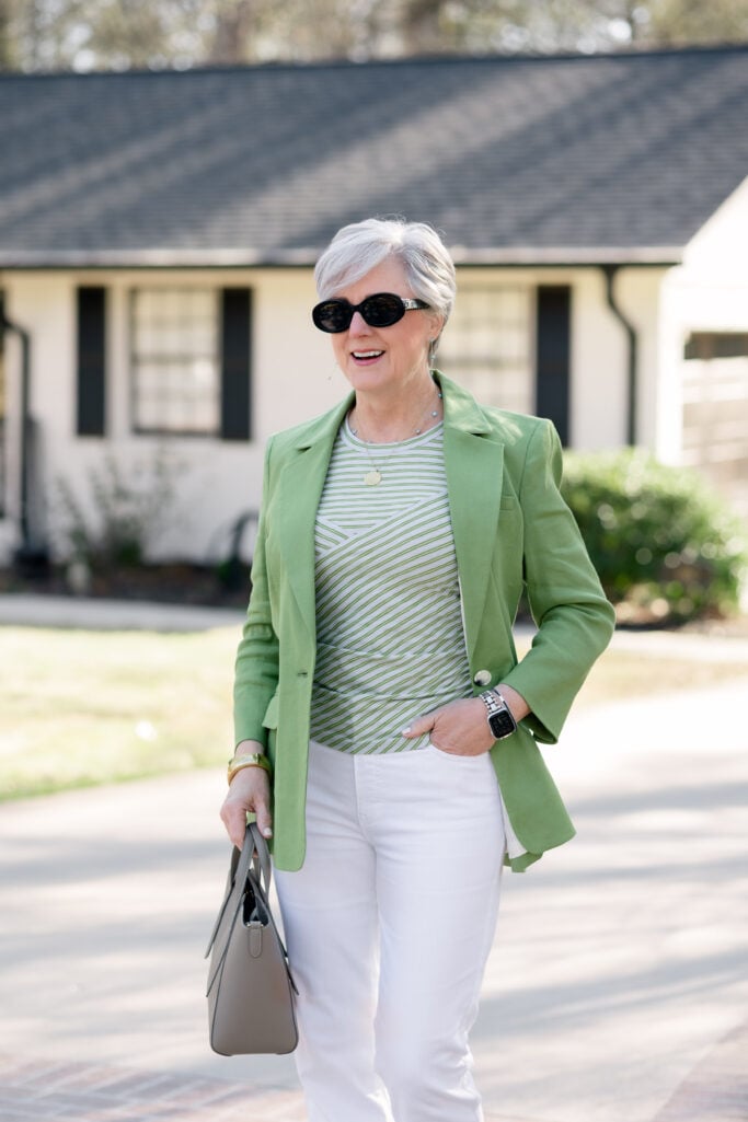 Close-up of woman over 50 with silver hair smiling in a bright green blazer over a green and white striped tee, white denim, oval black sunglasses, a gold pendant necklace, and a grey structured mini tote — showing how to wear spring green over 50 with a tonal stripe and clean white base.