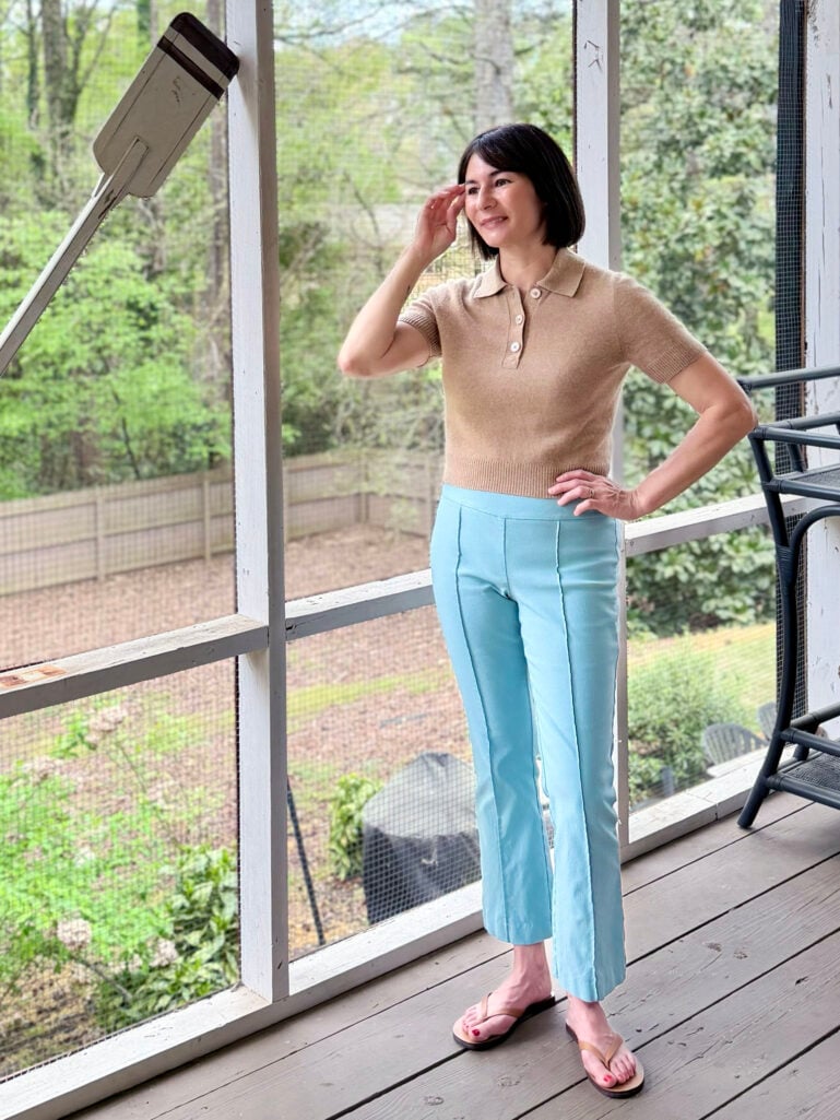 Kelly stands on the screened porch in a camel short-sleeve polo sweater and sky blue slim ankle trousers with a front seam detail, one hand raised to her hair, smiling over her shoulder.