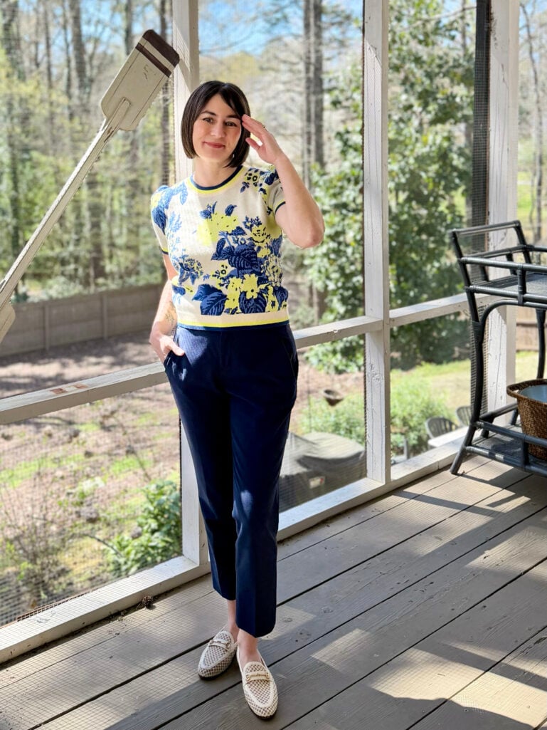 Kelly stands on the screened porch in a blue and yellow large-scale floral puff sleeve sweater tee and navy slim ankle pants, one hand raised to her hair, smiling softly at the camera.