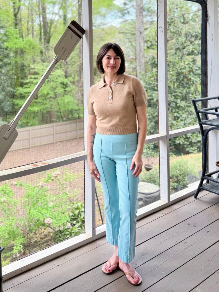 Full-length view of Kelly on the screened porch in a camel polo sweater, sky blue J.McLaughlin Ivy Pants, and easy summer flip-flops, standing relaxed with hands at her sides and smiling at the camera.