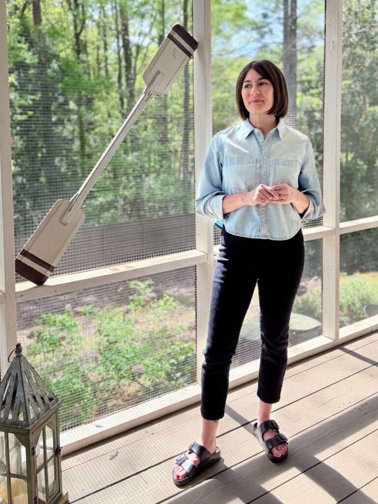 Full-length view of Kelly on the screened porch in her light chambray Étienne cropped top, black Derry Illusion Denim Pants, and black Arizona EVA Birkenstock sandals, leaning slightly and smiling at the camera.