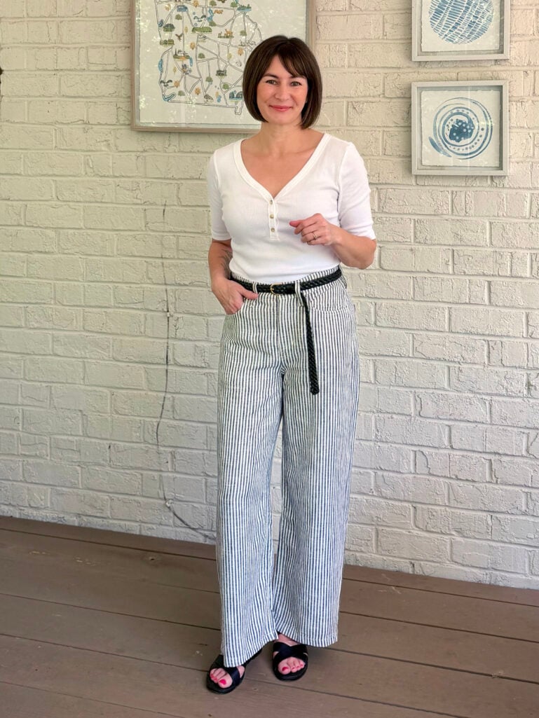 Full-length view of Kelly on the porch in her white ribbed henley, wide-leg seersucker stripe pants with black belt, and black slide sandals, one hand in her pocket, smiling at the camera.