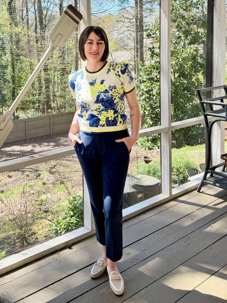 Kelly leans against the screened porch railing in her blue and yellow floral sweater tee and navy ankle pants with cream mesh bit loafers, both hands in her pockets, smiling at the camera.