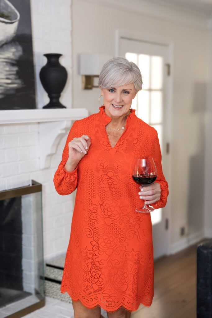 Woman over 50 with silver hair wearing a bold orange lace shift dress with a ruffled mock neckline and scalloped hem, holding a glass of red wine indoors — an example of how to wear orange over 50 with confidence for an evening occasion or entertaining at home.