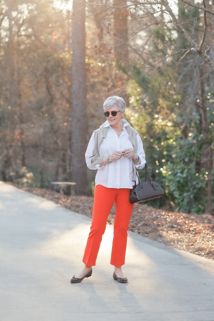 Woman over 50 with silver hair wearing bright orange flare ponte pants with a white button-down shirt, a neutral linen vest layered over the shirt, leopard print pointed-toe flats, and a black structured satchel bag — showing how to style orange pants for spring over 50 with a crisp neutral top.