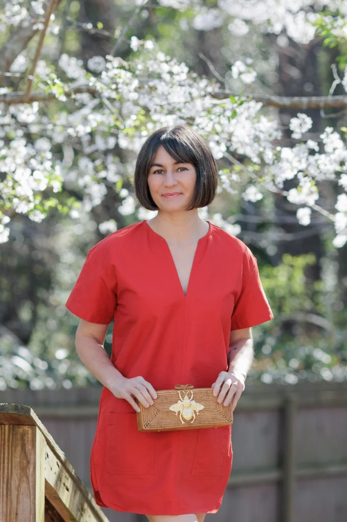 Woman wearing a rust-orange V-neck short-sleeve shift dress with patch pockets, holding a handwoven rattan box clutch with a gold bee detail, photographed outdoors against spring blossoms — a warm-toned spring look in orange-red for women over 50.