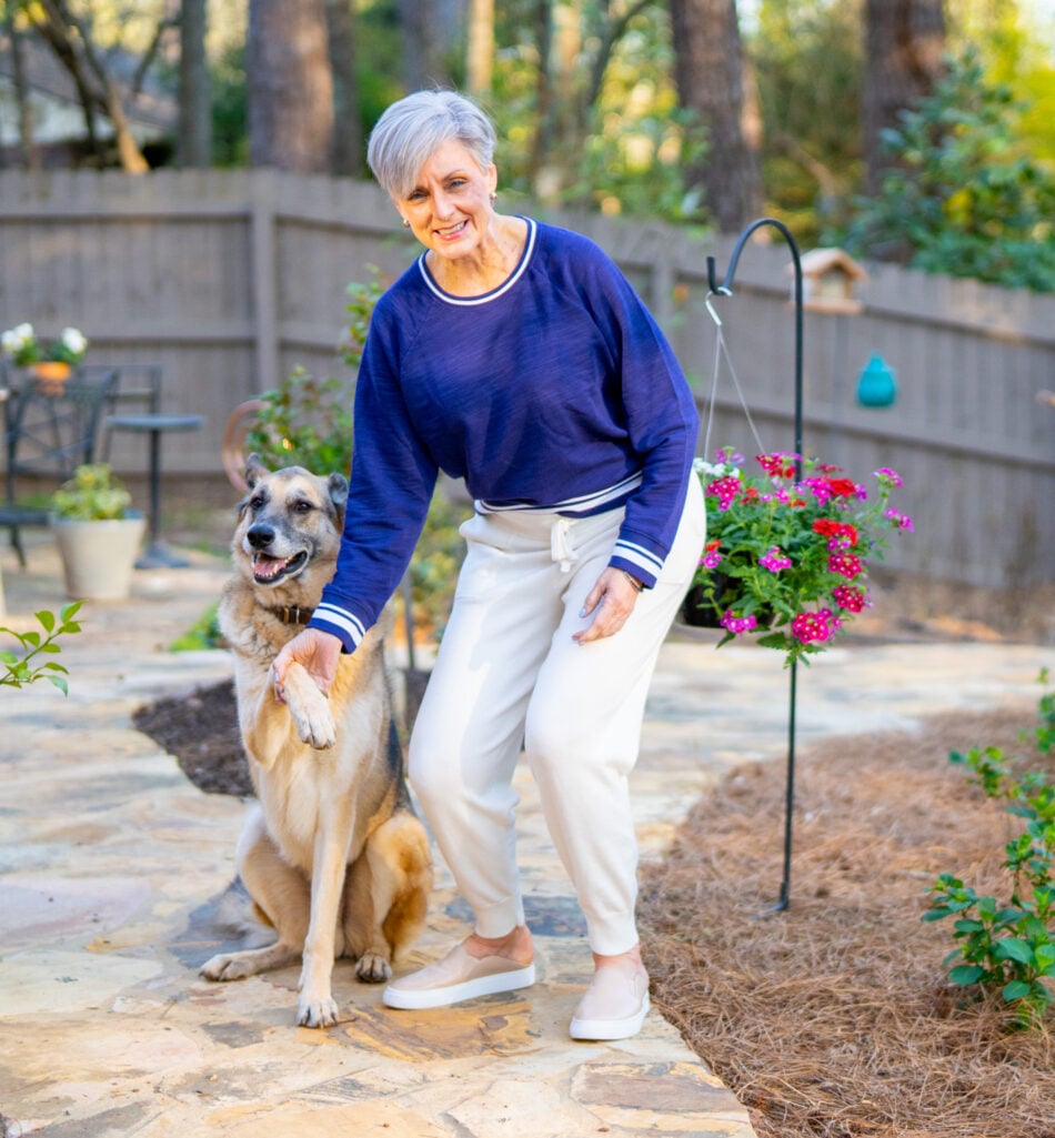 Beth stands outdoors on a stone garden patio in 2021, wearing a navy sweatshirt and cream pants, smiling as she holds Oscar's paw — Oscar, a large happy German Shepherd mix, sits beside her with a big smile, a hanging basket of pink and red verbena blooming in the background.