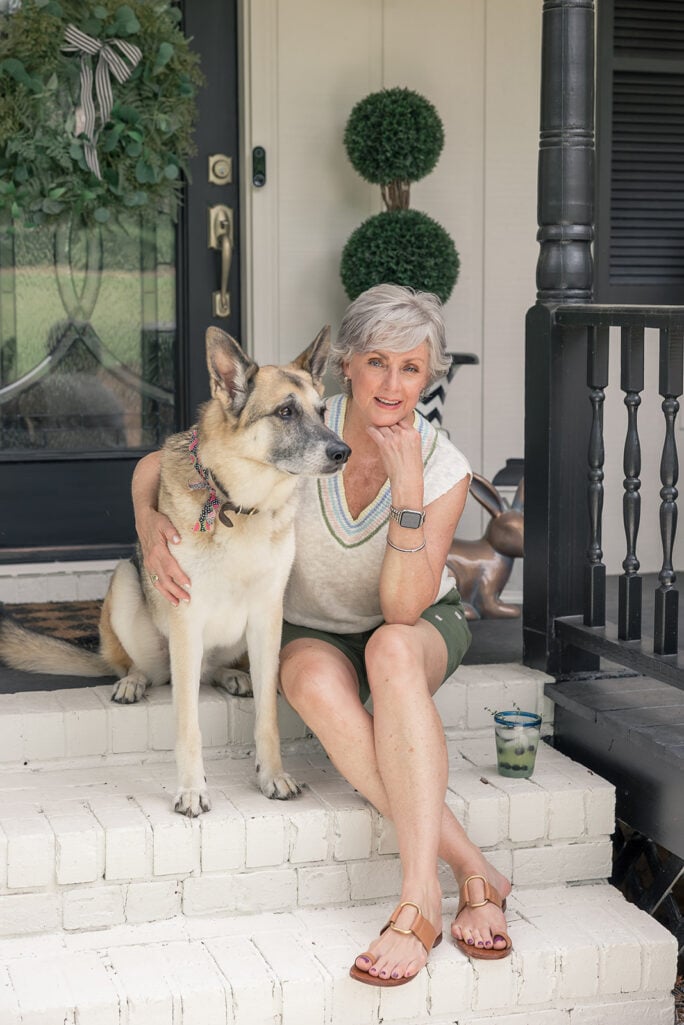 Beth sits on the white brick front porch steps in olive shorts and a striped sweater vest, her arm around Oscar, her German shepherd, who leans against her contentedly. Topiary ball trees and a front door wreath are visible behind them.