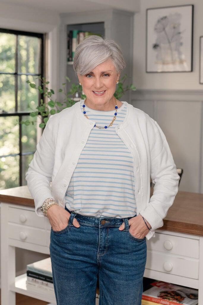 Woman over 50 with silver hair standing indoors in a gray-paneled study with a black-framed window and bookshelf behind her, wearing a white sweatshirt-style button-front cardigan layered over a blue and white horizontal striped crewneck tee, paired with medium wash straight-leg girlfriend jeans, a long gold chain necklace with cobalt blue and clear glass beads, gold pearl stud earrings, and stacked pastel and gold beaded bracelets — an example of styling a lightweight white cardigan as a spring layer over 50.