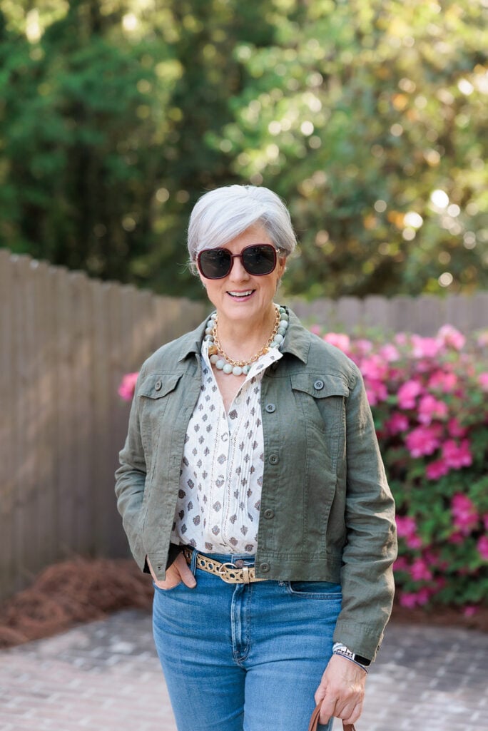 Woman over 50 with silver hair smiling outdoors in front of a wooden fence with bright pink azalea bushes in bloom, wearing dark tortoise oversized sunglasses, a chunky pale green and gold beaded statement necklace, a cream cotton-eyelet blouse with delicate medallion print, a cropped olive green linen button-front jacket with chest pockets, medium wash high-rise straight-leg jeans, and a woven gold metal belt — a structured cropped linen jacket styled as a spring layer for women over 50.