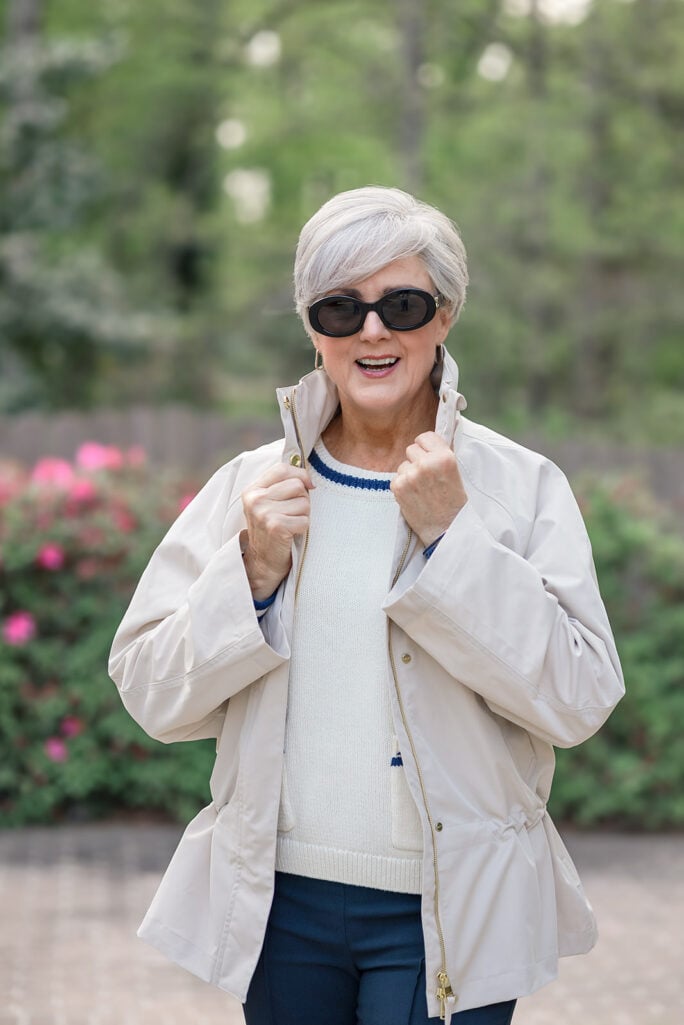 Woman over 50 with silver hair walking on a paved driveway with green trees and pink azalea bushes behind her, wearing dark oval Celine sunglasses, an ivory drawstring-waist lightweight rain jacket with a stand collar and gold zipper layered over an ivory crochet-textured crewneck sweater, navy slim ankle pants, and nude ballet flats — demonstrating how to layer a light rain jacket with a sweater and trousers for spring weather over 50.