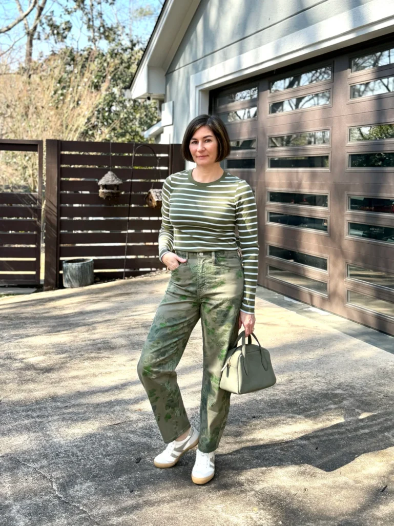 Woman wearing an olive green and white horizontal striped long-sleeve fitted tee tucked into wide-leg high-rise barrel jeans in an olive abstract print with green painterly detail, paired with white and tan Gola sneakers and a sage structured top-handle satchel bag — a sophisticated example of tonal pattern mixing that keeps two prints in the same color family for a polished, wearable look over 50.