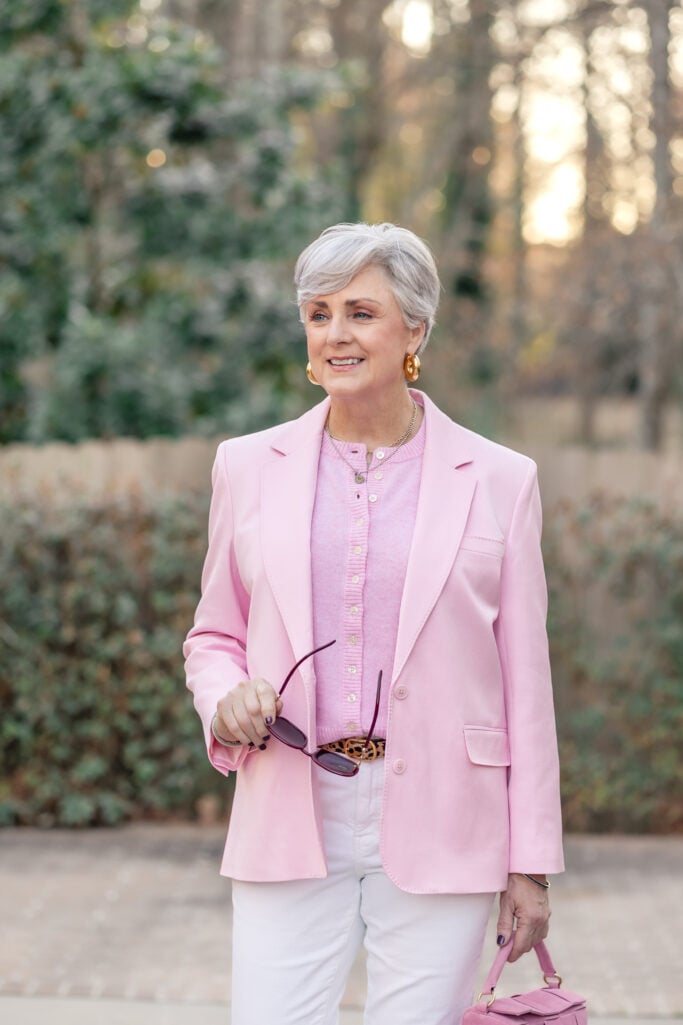Woman over 50 with silver hair wearing a tonal pink look — a structured pale pink blazer layered over a matching pink ribbed cardigan with gold buttons, white denim jeans, a leopard print belt, chunky gold hoop earrings, a layered gold necklace, and a small pink suede top-handle bag — a polished example of how to wear pink over 50 in spring 2026.