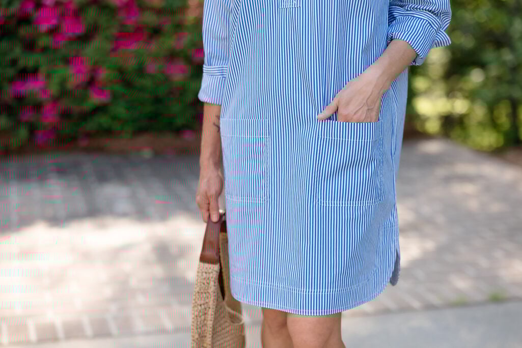 Close-up of blue and white striped broadcloth shirtdress showing front patch pocket and hem detail with straw bag — petite-friendly shirtdress features