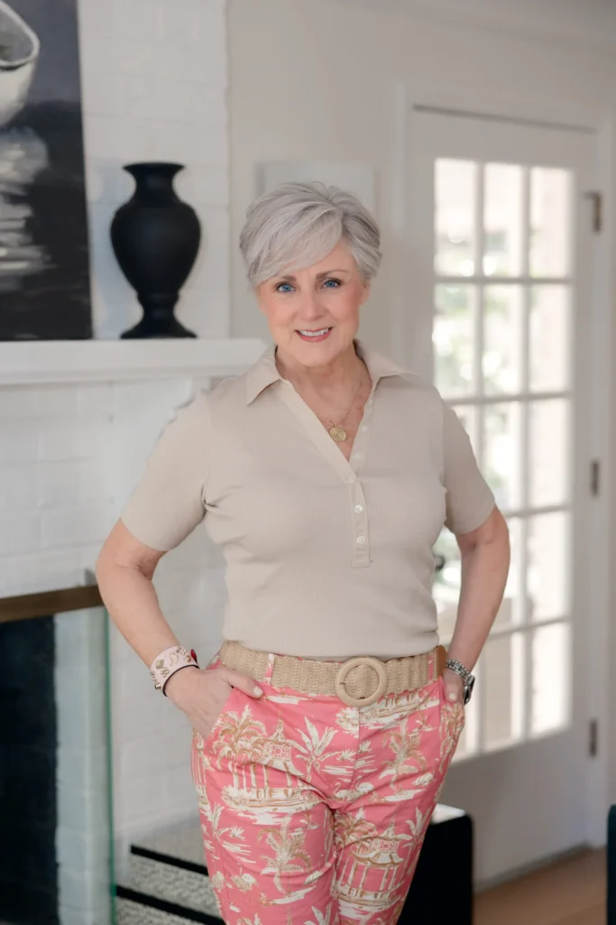Close-up of woman over 50 with silver hair wearing a beige ribbed short-sleeve polo top with a gold medallion pendant, layered pink and red beaded bracelets on one wrist and a silver watch on the other, paired with pink and tan palm-and-pagoda print straight-leg pants cinched with a natural woven raffia belt featuring a round woven buckle — a close-up styling of how a raffia belt adds texture to a spring print pant over 50.