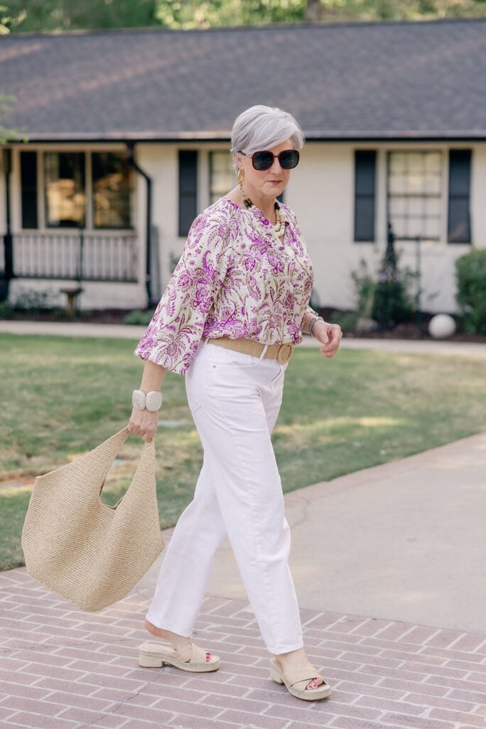 Woman over 50 with silver hair wearing dark tortoise sunglasses, gold wooden disc drop earrings, a wooden bead statement necklace, and a cream and magenta paisley print blouson top with three-quarter sleeves, paired with white straight-leg jeans cinched with a natural woven raffia belt with a round buckle, carrying an oversized natural raffia slouchy shoulder bag, and wearing crisscross woven raffia block heel slide sandals with espadrille soles — a vibrant, print-forward raffia styling for spring.