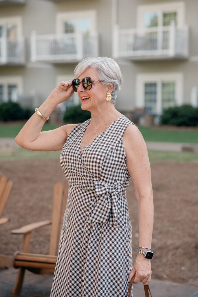 Close-up of woman over 50 with silver hair laughing outdoors wearing a black and white gingham wrap midi dress with a tie waist, oversized tortoise and gold square sunglasses, large gold sculptural drop earrings, and a gold bangle bracelet — a detail shot showcasing sunglasses and jewelry as Mother's Day gift ideas for women over 50.