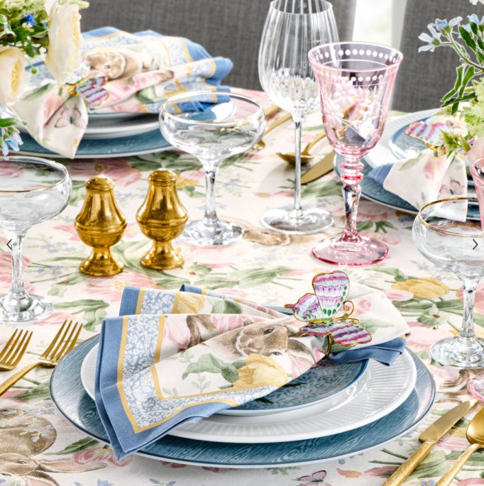 Colorful spring tablescape with floral tablecloth, layered blue and white plates, gold flatware, pastel glassware, and patterned napkins with bunny and butterfly accents.