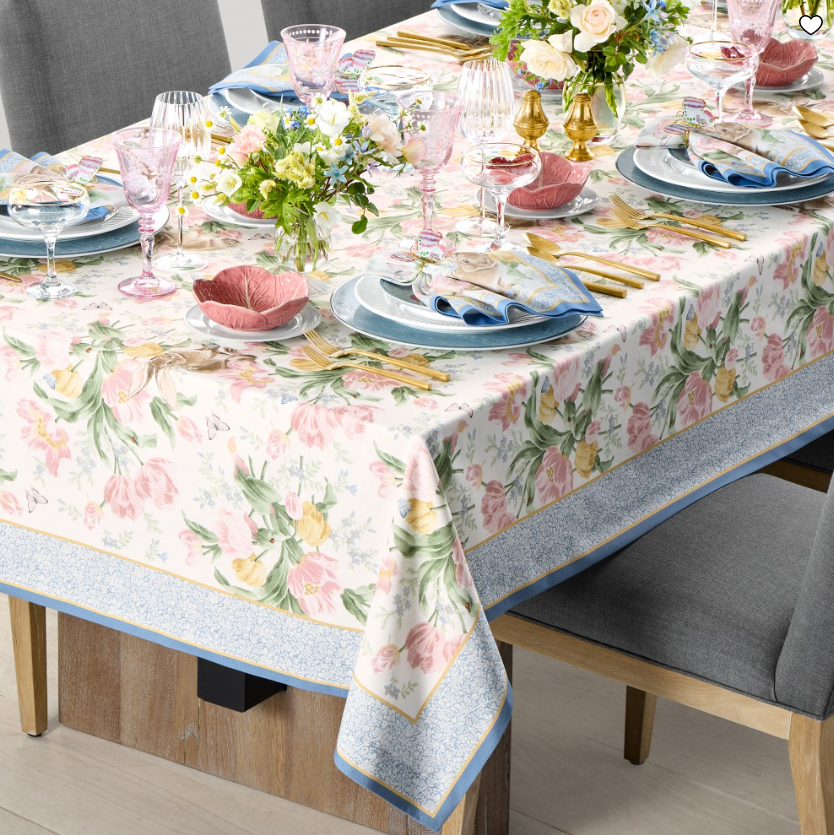 Floral garden tablecloth with soft pastel blooms in pink, yellow, and green, styled on a dining table with layered place settings, gold flatware, blue napkins, and fresh spring floral arrangements.