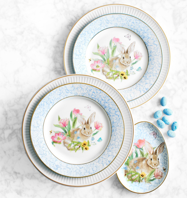 Spring-inspired dinnerware set with blue and white patterned plates featuring a bunny and floral design, accented with gold rims and pastel details.