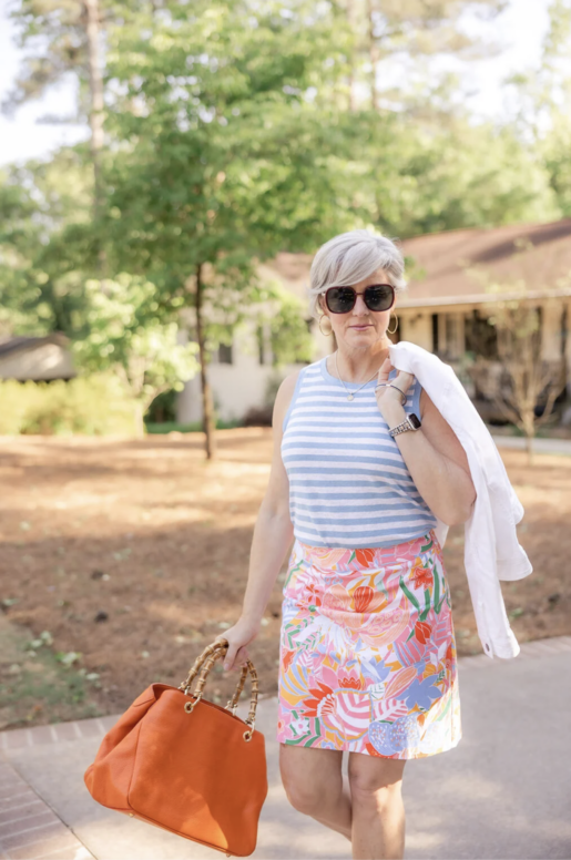 Woman with short gray hair wearing a blue striped sleeveless top and colorful floral skirt, carrying an orange handbag and white jacket while walking outdoors on a sunny day.